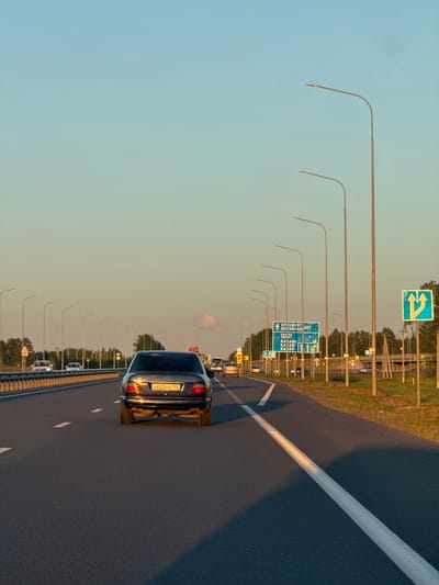 The image shows a view from inside a moving vehicle on a highway in the vicinity of Bol'shie Kabany, Russia, during the late afternoon or early evening. The sky is a clear gradient of light blue to pale yellow. Tall, modern streetlights line the right side of the highway. A dark-colored sedan with the license plate A622HO 155 is prominently visible in the foreground, traveling in the same direction. Several other vehicles are present on the highway, including cars and what appears to be a bus in the distance. Road signs are visible on the right side of the highway, including a blue directional sign indicating routes to Orenburg, SOKRY, KAZAN', and KAZAN' with distances, and a blue and yellow traffic sign with arrows indicating lane changes. Trees and foliage line the sides of the highway. The road surface is asphalt with visible white lane markings. A long shadow stretches across the right lane from the perspective of the viewer's vehicle.