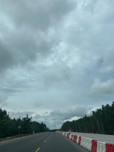 The image depicts a two-lane asphalt road under a cloudy sky. The road is lined with red and white plastic barriers on the right side, indicating ongoing construction or a divided lane. Dense green trees form a border along both sides of the road. A triangular road sign is visible on the left side of the road, partially obscured by trees. The scene appears to be in a rural or semi-rural area, likely a roadside construction site. The overall atmosphere is overcast and possibly damp.