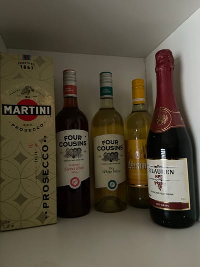 A cluttered shelf in a white cabinet contains several bottles of alcoholic beverages and a box.
At the top is a bottle of "St. Lauren Red Sparkling Fruit Drink, Red Grape". Beneath this is a bottle labeled "49 STR" and "Sweet White Wine." Below this is a bottle of "Four Cousins Dry White Wine." Next is a bottle of "Four Cousins Natural Sweet Rosé Wine." At the bottom is a partially visible box of "Martini Prosecco."
There are no visible indications of Isheri, Nigeria or people in the image.