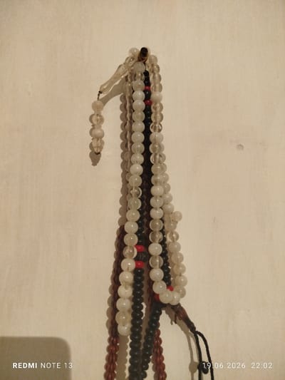A close-up, eye-level shot shows multiple strands of prayer beads hanging vertically against a textured, cream-colored wall. The beads vary in color and material, including translucent white, opaque white, black, and reddish-brown. Some strands are composed of larger, round beads, while others have smaller, more elongated beads. There are also red beads interspersed on some strands. A dark tassel is visible at the bottom of one strand. A shadow of an unseen object is cast on the wall in the lower left corner, and the text "REDMI NOTE 13" is present in the bottom left, alongside a timestamp "19.06.2026 22:02" in the bottom right. The lighting is dim, casting shadows from the beads onto the wall. The scene appears to be indoors, likely in a residential setting. There are no people or animals present in the frame. The location is identified as Асан-Чек, Kyrgyzstan, though the visual elements do not provide specific indicators of this geographical location beyond the provided metadata.
