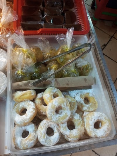 The image captures a close-up, high-angle view of various food items displayed for sale at a market stall in Lê Thanh Nghị Ward, Vietnam. In the upper segment, a red plastic basket holds multiple dark brown, individually plastic-wrapped confectionery items, predominantly rectangular with some semi-circular edges. Below this, a white translucent plastic tray features two distinct layers of food. The upper layer contains numerous small, round, yellowish-green items, each individually sealed in a clear plastic bag tied with a yellow closure. A pair of silver metal serving tongs rests diagonally across these bagged items. A horizontal brown cardboard tube or rolled paper acts as a divider, separating the bagged items from the lower section of the tray. This lower section contains approximately 15-20 light-golden, ring-shaped donuts, generously coated with white powdered sugar. The overall display rests on a metallic surface, with a blurred reddish-orange object and a light-colored patterned floor visible in the background.