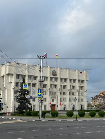 The image shows a building in the city of Akhieli, Georgia. The building is white and has many windows. There are flags flying on the building's roof, including the flag of Georgia and another flag with red, yellow, and white stripes. The building is located next to a street with trees and bushes along the sidewalk. There are several traffic signs visible, including a pedestrian crossing sign. The sky is cloudy.