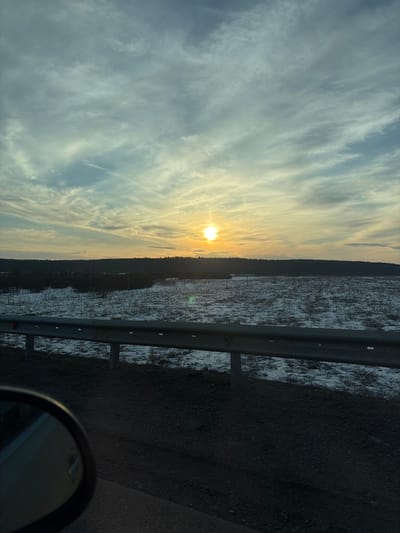 The image shows a view from a vehicle traveling on a road near Chernovskoye, Russia. The sky is predominantly blue with wispy white clouds tinged with yellow from the setting sun, which is low on the horizon. A dark treeline separates the sky from a snow-covered field, visible on the right side of the frame. A metal guardrail runs along the road, and the dark asphalt surface is prominent. A portion of the vehicle's side mirror is visible in the lower right corner. There are no people visible in the frame.