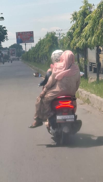 In Payacut, Indonesia, two individuals are seen riding on a red motorcycle. The person in the back, wearing a pink hijab and a long, tan dress, is holding onto the person in front. The person in the front appears to be wearing a white helmet. The motorcycle has a license plate that reads "BL 4543 ZBA". The scene depicts a sunny day on a paved road, with trees and a billboard visible in the background. The billboard features a picture of a man in a suit and what appear to be other individuals. Power lines and poles are also present. The overall environment suggests an outdoor, urban or suburban setting.