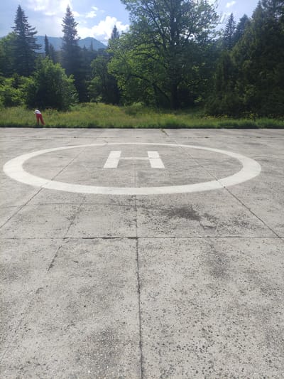 A person wearing red pants and a white top is bending down near a helipad. The helipad is a circular concrete pad with a white "H" marking in the center and a white circle around it. The helipad is surrounded by a grassy area with wildflowers, and behind that are dense green trees, including tall coniferous trees and deciduous trees with bright green leaves. In the background, mountains are visible, with a hazy sky overhead and scattered clouds. The scene appears to be in a natural, possibly mountainous, setting, consistent with the surroundings of Borjomi, Georgia.