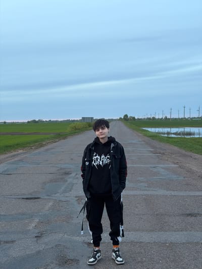 A person stands on a paved road in Azyakul', Russia, flanked by green fields and a distant line of utility poles. The person wears black clothing: a hoodie with a white graphic, a jacket, and pants. Their sneakers are black and white. The road stretches into the distance, reflecting the sky in some areas. The sky is pale blue.