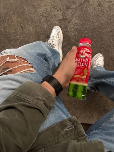 An individual is seated outdoors, their legs clad in ripped blue jeans and white sneakers. The person holds a red bottle of OKF Watermelon drink with aloe. The background consists of a textured, dark grey ground, likely concrete, common in urban settings. The presence of this type of paving and the general appearance of the environment suggest an outdoor public space. While the image depicts a common urban scene, there are no specific visual cues that definitively place the location within the city of Nantes, France.