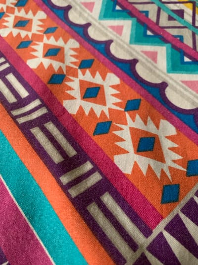 This media file shows a close-up, abstract view of a colorful fabric with a geometric pattern. The fabric is draped, creating folds and showcasing the texture of the material. The dominant colors are vibrant orange, pink, purple, teal, and white, arranged in diagonal bands and geometric shapes. These shapes include diamond-like figures, zig-zag patterns, and striped elements. The lighting is consistent across the image, suggesting an indoor setting or an overcast day, with no distinct shadows indicating a specific time of day. There are no people, animals, or any clear indication of a specific setting or location beyond the fabric itself. The image focuses solely on the visual details of the textile's design and texture. No text is visible in the image.