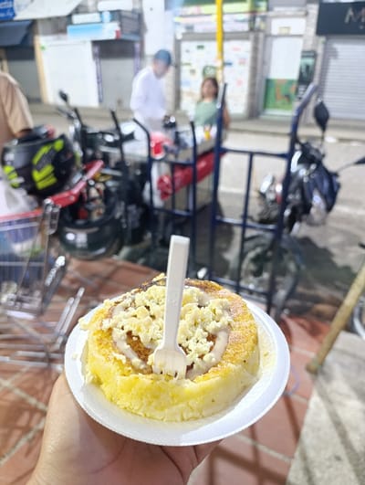 A hand holds a white plate with a cylindrical cake. The cake is yellow, topped with white crumbs and a spiral of white icing, held in place by a white plastic fork. In the background, out-of-focus, are various motorcycles, a shopping cart, and two people standing near what appears to be a food cart or stand, set on a bricked area. The scene is lit unevenly, suggesting an outdoor setting at dusk or night.