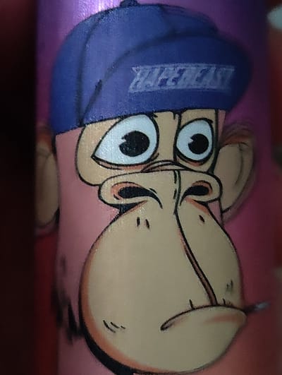 The image is a close-up, head-and-shoulders shot of a cartoon drawing of a monkey wearing a blue baseball cap. The monkey has large, cartoonish eyes, a pink face, and a light brown nose and mouth area. The cap has white lettering on it, which appears to say "HYPEBEAST." The background is a blurred, textured purple and pink. The drawing is on an object, possibly a cylindrical container, given the curvature of the face. There is no visible indication of the city of Dorst, Netherlands, or any urban environment.
