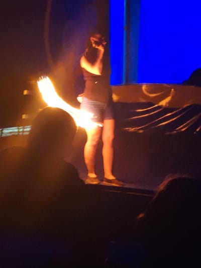 The image depicts a fire performance taking place indoors, likely on a stage, viewed from the audience's perspective. The setting is predominantly dark, with selective lighting highlighting the performer and parts of the background.
At the center of the image, a person, presumably the performer, stands facing slightly towards the right of the frame. They are wearing a dark sleeveless top or tank top and light-colored shorts, possibly denim. Their skin is illuminated by a warm, orange-yellow light emanating from a fire prop they are holding with their left hand. This prop generates a large, bright flame that curves upwards and outwards to the performer's left, appearing as multiple distinct streaks of fire, suggesting a multi-pronged torch or a fire fan. The performer's right arm is bent, and their hand is near their mouth or holding an object, possibly a microphone. Their legs are visible, wearing dark, casual slip-on shoes or sneakers.
Behind the performer, to the right, a prominent vertical section of bright blue light is visible, possibly from a screen, light panel, or stage backdrop. Below this blue light and behind the performer, there is a dark surface with some light-colored, draped fabric or material that catches some of the warm light, creating wavy patterns.
In the foreground, silhouetted heads of two audience members are visible. One is in the lower-left corner, and another is in the lower-right corner, both facing towards the stage and the performer. They are out of focus, emphasizing the performer and the fire.
The overall atmosphere is theatrical and dramatic due to the stark contrast between the intense, bright fire and the surrounding darkness. The activity is clearly a fire show or performance, characterized by the active manipulation of flames by a performer. The time of day is not explicitly stated but implied to be evening or night due to the dark environment and stage lighting. No text is visible in the image.