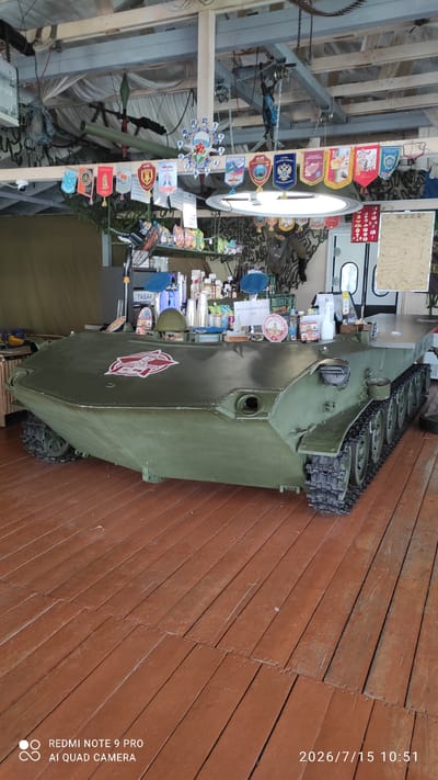 The image shows a bar counter made from a green, tracked military vehicle, likely an armored personnel carrier. The vehicle is positioned indoors on a wooden floor. The top of the vehicle has been flattened and covered to serve as a bar surface, displaying various bottles, cups, a military helmet, and signage. Several small flags and banners, many with emblems and text, are hung above the bar area, suggesting a themed establishment possibly related to military history or patriotism. A clock with a distinctive design featuring a star and wings is also visible. The background reveals more decorations and possibly seating areas. A sign with the Cyrillic text "ТАБАК" (tobacco) is visible on a pole behind the bar. The overall setting appears to be a casual, possibly outdoor or semi-outdoor, venue. There are no people clearly visible in the scene.