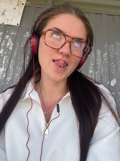 A young woman with long, dark brown hair is facing the camera, her head tilted to the left. She has fair skin, brown eyes, and is wearing tortoise-shell framed glasses. She is sticking her tongue out slightly. Red headphones are around her neck, connected to wires that lead to her white, zippered jacket. A silver necklace with a cross pendant is visible. The background appears to be a weathered, gray surface with peeling paint, possibly an exterior wall. The location is Molokovo, Russia.