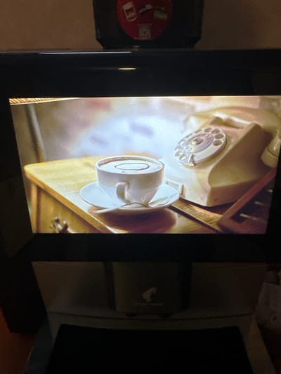 The image shows a close-up view of a vintage rotary telephone and a cup of coffee on a wooden table. The telephone is beige and has a circular dial with numbers. The coffee cup is white and filled with a dark liquid, possibly hot chocolate or coffee, with a swirl pattern on top. A saucer and a spoon are also visible. The setting appears to be indoors, with soft lighting. There is no indication of the location being in Vama Veche, Romania, nor are there any people or specific actions depicted. The image is framed by the edges of a screen.