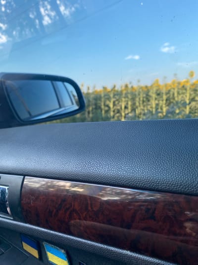 An interior shot of a car shows a driver's side door panel with a wood-grain accent and a rearview mirror reflecting a field of sunflowers under a bright blue sky with scattered clouds. The field of sunflowers is blurred in the background. Two patches resembling the Ukrainian flag are visible on the lower part of the door panel. The image is taken from a fixed perspective, suggesting the car is stationary. The location is identified as Maiske, Ukraine.