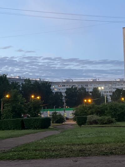 The image depicts a residential area in Rahya, Russia, at dusk. A long, multi-story apartment building dominates the background, its windows dimly lit. In front of the building, a dense line of dark green trees partially obscures the lower floors. Several streetlights with bright yellow lamps are visible among the trees, casting an artificial glow on the surroundings. In the foreground, a paved path curves through a grassy area with neatly trimmed hedges. The sky above is a pale blue with wispy, elongated clouds. The overall scene suggests a quiet evening in a suburban or semi-urban environment.