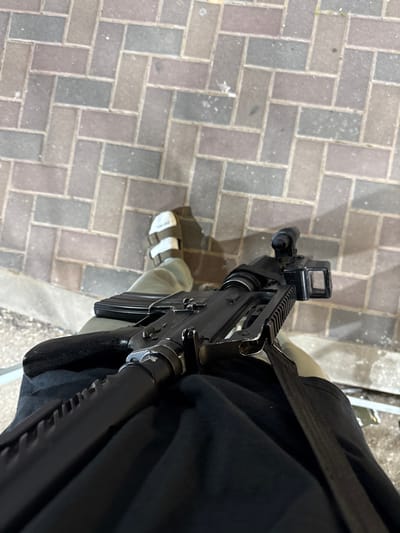 The image shows a black rifle lying across the lap of a person. The person is wearing a black shirt, green camouflage pants, and a pair of white sandals. The person is standing on a paved sidewalk made of brick. The rifle is resting on the person's lap, pointing to the left. The rifle has a scope and a black sling attached to it. The image is taken from a high angle, looking down on the person and the rifle. The image was likely taken in Mazkeret Batya, Israel.