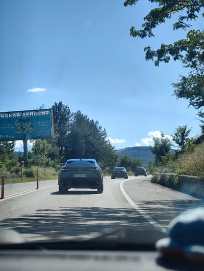 A car's-eye view from a vehicle traveling on a road in Malinovo, Bulgaria, on a sunny day. The road curves to the right, bordered by a concrete retaining wall on the right and trees and vegetation on both sides. Several other cars are visible ahead, moving in the same direction. In the distance, rolling hills and more trees are seen under a clear blue sky with scattered clouds. A billboard advertising a bank deposit is visible on the left side of the road, near the beginning of the curve. The interior of the vehicle, including a blurry view of a blue hat in the foreground, is also captured.