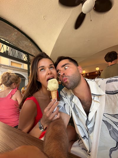 A young Caucasian couple is posing for a selfie while eating ice cream cones. The woman on the left is wearing a red top and holding a vanilla ice cream cone. She is looking up with her mouth open, about to take a bite of the ice cream. The man on the right is wearing a white shirt with a black floral pattern and is puckering his lips, making a funny face. He is leaning towards the woman, and his eyes are wide with a playful expression. In the background, out of focus, a woman in a pink top is visible. The scene appears to be outdoors, possibly at a cafe or restaurant, with a ceiling fan visible overhead. The background suggests an urban setting with buildings and architectural details. The image is taken from a low angle, emphasizing the subjects.