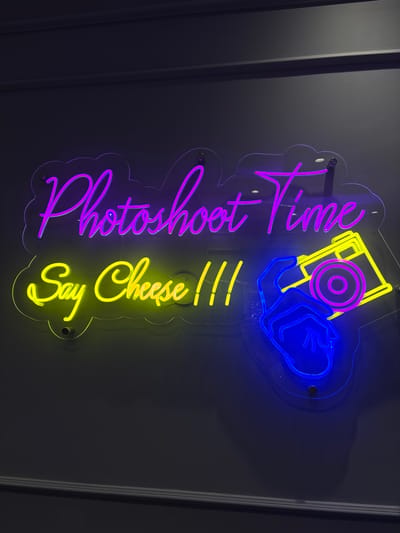 A neon sign with the words "Photoshoot Time" in purple and "Say Cheese !!!" in yellow is mounted on a dark gray wall. To the right of the text, a neon illustration depicts a blue hand holding a yellow camera with a purple lens and body. The sign is supported by clear acrylic backing and secured with metal screws. The environment appears to be an indoor space, likely a studio or a themed room, in Sweileh, Jordan.