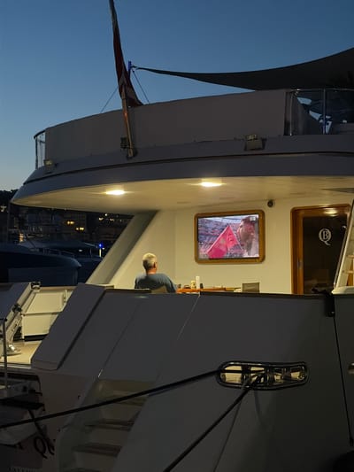 An exterior shot at dusk shows a person seated in front of a television on the deck of a yacht docked in Imperia, Italy. The person, a male with short gray hair, is wearing a blue shirt and is facing away from the camera, looking at the television. The television displays a sports broadcast, with a person holding a pink flag in the foreground. The yacht has a white hull and upper structure, with a dark-colored canopy overhead. In the background, other yachts are visible, as well as buildings with illuminated windows along the waterfront. The sky is a deep blue, indicating twilight. The overall scene suggests a relaxing evening on the water.