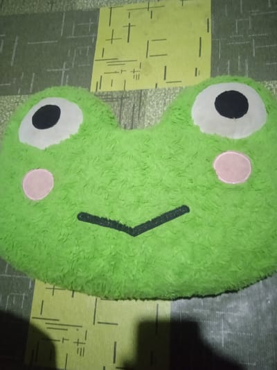 A green plush pillow shaped like a frog's head rests on a surface with a patterned fabric. The pillow has large white eyes with black pupils, and two pink circles on its cheeks. A black line forms its mouth. The patterned surface beneath the pillow consists of alternating green and yellow rectangular sections with thin white lines and some black markings. The image is taken from a slightly overhead angle, with the pillow dominating the frame. The lighting appears to be dim, casting some shadows. The specific location within Bora, Indonesia, is not ascertainable from the visual information provided.