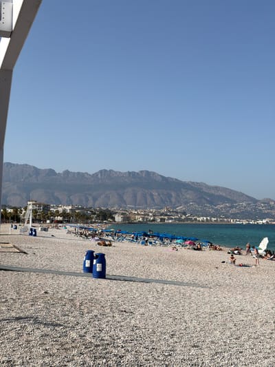 The image depicts a broad, pebble-covered beach in Pla Parcial Platja, Spain, under a clear blue sky. In the foreground, two blue trash receptacles stand on the pebbled ground, with a shaded walkway leading past them. Beyond the receptacles, the beach is populated with numerous beachgoers, many relaxing on lounge chairs under blue and pink umbrellas. A few individuals are visible wading in the shallow, turquoise water. In the middle ground, a line of buildings, including a prominent white lifeguard tower, stretches along the coastline, interspersed with palm trees. The background is dominated by a majestic mountain range with rugged peaks, forming a dramatic backdrop to the coastal scene.