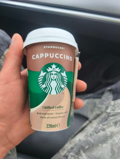 A hand holds a Starbucks "CAPPUCCINO" chilled coffee cup. The cup is brown with a white lid and features the Starbucks siren logo in green. Text on the cup describes the drink as "Bold espresso," "Creamy milk," and "Hints of cocoa flavour." The volume is listed as "220ml e." A wooden stirrer is attached to the side of the cup. The background is blurred, showing what appears to be the interior of a vehicle, with a glimpse of camouflage fabric. The location is identified as Unknown, Ireland.