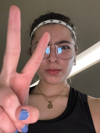 The image displays a close-up, chest-up portrait of a single female individual in an indoor setting within Komchén, Mexico. The subject has dark hair pulled back from her face, secured by a white athletic headband adorned with repeating black Adidas logos. She wears round, metallic-rimmed eyeglasses, with reflections of bright, rectangular light sources visible in the lenses. Freckles are present across her nose and cheeks. Her facial expression includes a furrowed brow and a direct gaze towards the camera, with her left eye (viewer's right) appearing slightly wider than her right eye. She is dressed in a black tank top or athletic garment, accessorized with a thin gold chain necklace featuring a circular gold pendant with an emblem, and a small gold hoop earring in her right ear. Her left hand is raised prominently in the foreground, forming a "peace sign" gesture with the index and middle fingers extended. The nails on these extended fingers are painted a light blue color. The background is a dim, indistinct indoor environment, featuring blurred lighter horizontal and diagonal shapes.