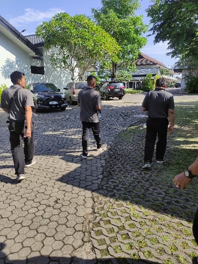 The image shows three men walking on a cobblestone path outside of a white building. The men are all wearing dark clothing. The path is bordered by green grass. Two cars are parked near the path and the building. The image was taken in Gambiran, Indonesia, at coordinates: -8.434, 114.18. A blue sky with white clouds is visible in the background. The man closest to the camera is walking towards the right, the other two men are walking towards the left of the frame.