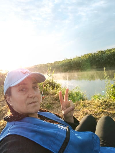 The image shows a woman wearing a blue vest and a light blue baseball cap, sitting by a river. She is making a peace sign with her right hand. Two fishing rods are visible in the background, planted in the ground near the riverbank. The sun is setting or rising, casting a warm glow over the scene. The river is calm, with a light mist rising from its surface, and lush greenery lines the opposite bank. The ground where the woman is sitting appears to be covered in dry grass. The location is Dzhiginka, Russia.