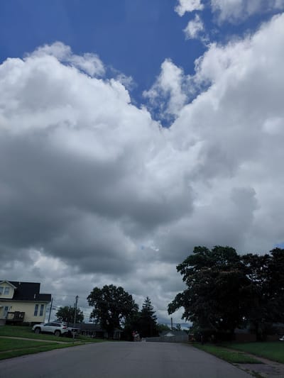 This media file captures an outdoor scene in Nicholasville, United States of America, likely during the daytime under cloudy skies. The image is oriented vertically, with the sky dominating the frame. Large, fluffy white clouds contrast with darker, more ominous gray clouds, suggesting a dynamic weather system. A patch of clear blue sky is visible on the left.
In the lower portion of the image, a residential street is visible, with a paved road curving to the right. Several houses with pitched roofs are present, one painted yellow with dark trim and a porch. A white SUV is parked on the grass in front of one of the houses. A large, dark green tree with dense foliage is prominent on the right side of the frame, casting shadows. Power lines and poles are also visible, indicating a developed area. The overall mood is somewhat subdued due to the overcast conditions. No people are visible in the image, and no text is present.