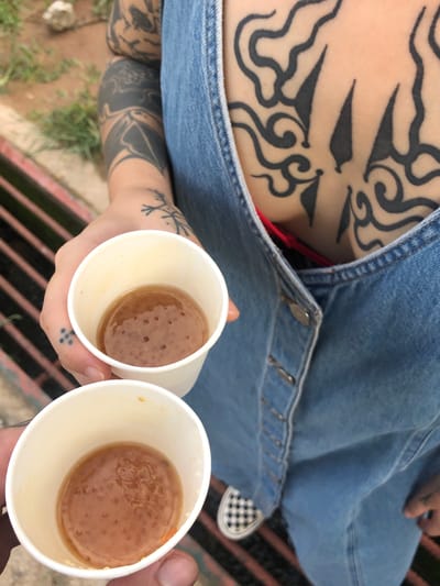 A person with extensive tattoos holds two white paper cups filled with a brown, pulpy liquid, likely a beverage containing tapioca pearls. The person is wearing a light blue denim overall dress. Visible underneath the dress neckline is a red garment. The person has tattoos on their chest and arms. Their left hand has several dot tattoos between the thumb and forefinger, and there is a tattoo of a sprig on the left forearm. Part of a black and white checkered shoe is visible, along with a metal grate underneath the individual. The setting appears to be outdoors, with vegetation and some kind of ground visible. There is no indication that this photo was taken in Pilana, Sri Lanka.