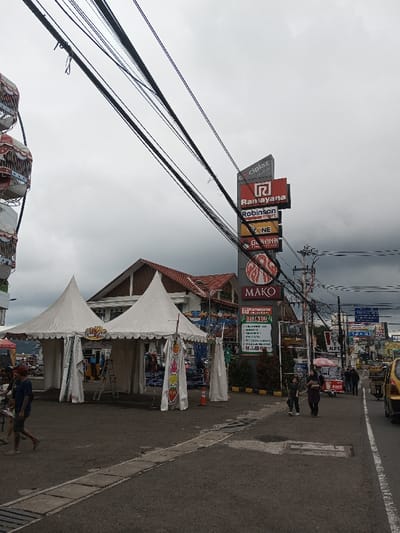 An outdoor market is underway in a city street. Two white tents stand to the left of the frame, near a tall sign advertising various businesses, including "Ramayana" and "MAKO." Utility poles and wires crisscross the sky, adding to the urban feel. People walk along the sidewalk, and a taxi idles in the street. The sky is overcast, suggesting a cloudy day.