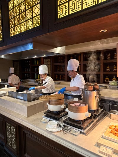 The image depicts an interior scene within a professional kitchen or buffet area in Tongxiang, China. Three individuals, dressed in white chef coats and tall white chef hats, are actively engaged in food preparation and service. Two chefs in the foreground wear blue disposable gloves and clear face shields.

The chef on the right is positioned near a large, cylindrical stainless steel multi-tier steamer emitting a prominent plume of white steam, indicating active cooking. To their left, two smaller ceramic pots with bamboo steamers are heating on individual gas burners; one steamer contains several pink/purple-hued food items, possibly taro cakes or buns.

Further left, a second chef is working at a station featuring stainless steel deep fryers, where metal baskets are visible. A third chef is partially visible in the far left background, facing away from the camera, working at another counter.

The serving counter in the foreground is light-colored, possibly marble or stone, and features a white cup, saucer, and tongs, along with small acrylic signs with Chinese characters. A tray of golden-brown baked goods or pastries is visible on the far right.

The background features dark wooden cabinetry and shelving units, displaying numerous glass jars filled with colorful ingredients, suggesting spices or preserved foods. The upper portion of the wall showcases ornate wooden panels with intricate gold-colored geometric latticework, characteristic of traditional Chinese design. A stainless steel ventilation hood hangs above the cooking area.