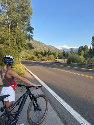 The image displays a female individual and a bicycle positioned on the paved shoulder of a two-lane road in a mountainous environment, identified as the vicinity of Aspen, Colorado, United States. The individual, observed from behind, possesses blonde hair styled in a ponytail and wears a white and blue bicycle helmet, a dark grey tank top, white shorts, and a red fanny pack/sling bag across her chest. Her left hand is resting on the handlebars of a dark blue Orbea Rise mountain e-bike, featuring a visible front wheel with disc brakes and tires marked "Wicked Will." The asphalt road exhibits a solid white line defining the shoulder and double yellow lines indicating the center. A metal guardrail parallels the road on the right side in the mid-ground. The immediate environment includes dense green foliage, with tall deciduous trees on the left and a variety of bushes and smaller trees lining both sides of the road. In the mid-distance, a brown wooden structure with a prominent stone chimney is visible on the right. The background is characterized by large, tree-covered mountains, with lighter, possibly snow-capped peaks and scattered white clouds visible in the far distance beneath a clear blue sky. The scene is illuminated by natural daylight, casting shadows on the road surface.