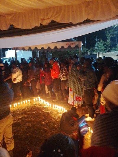 The image depicts a nighttime gathering in Marenyo-Shianda ward, Kenya, occurring under a large, light-colored fabric canopy structure. A substantial group of individuals, primarily adults, are present, standing and kneeling on a dark ground surface, likely dirt or short grass. The scene's primary illumination originates from numerous lit white pillar candles arranged on the ground, forming an elongated, angular pattern in the foreground and midground. Attendees are dressed in various casual civilian attire, including jackets, sweaters, and patterned garments. In the background, additional smaller tents are visible, one displaying the partial text "PCLANDA." Trees and distant structures are discernible against the dark sky. Specific actions include individuals observing the candle display, and one person in the foreground kneeling near the candles while holding an illuminated smartphone. The overall atmosphere indicates a communal event, possibly ceremonial or a vigil.