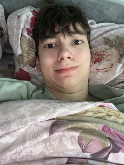This image depicts a young male individual lying in a bed in Zainsk, Russia. The subject, a young adult with fair skin, short dark brown hair, and dark brown eyes, gazes directly into the camera with a slight, closed-mouth smile. His chin is tucked, creating a visible fold of skin under his jawline. He is wearing a light green t-shirt, visible around his neck and shoulders. The individual is largely covered by patterned bedding, which features large floral designs in shades of cream, beige, pink, red, and purple on a light background. A lighter, possibly grey or white, pillow or headboard is visible behind his head. The scene is illuminated by ambient light, suggesting an indoor bedroom environment.