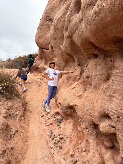 The image shows a woman in the foreground, standing on a dirt path next to a large rock formation. She is wearing a white t-shirt, blue leggings, and sneakers, and is smiling at the camera. In the background, two other people are walking up the path. The environment appears to be a desert or canyon area, with dry vegetation and red rock formations. The sky is overcast. Based on visual cues, the location is likely O'Neill Regional Park in Lake Forest, California, United States of America, known for its extensive network of hiking trails and sandstone canyons.