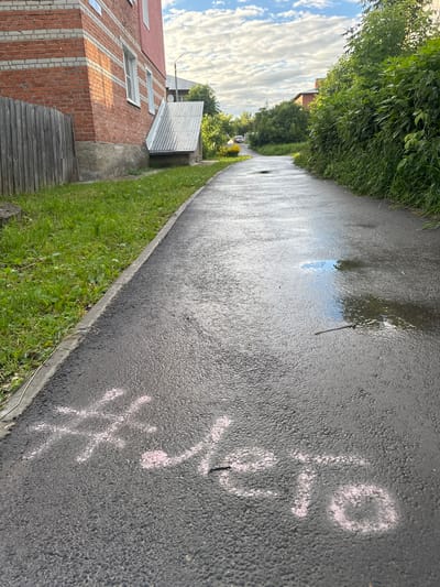 The image depicts a wet asphalt path with some pink chalk markings on it, including what appears to be a # symbol and some unreadable script. The path is bordered by lush green grass on one side and a paved area with a low concrete curb. To the left of the path, there is a brick building under construction with a corrugated metal roof attached to one side. Beyond the building, a dense line of trees and foliage forms a backdrop. In the distance, a car is visible on the road. The sky is partly cloudy with white clouds against a blue background. The presence of puddles on the path suggests recent rain, and the sunlight indicates it is daytime. The scene appears to be in a residential or semi-rural area, given the construction and greenery. The chalk markings are the most prominent detail on the path, but their meaning is unclear.