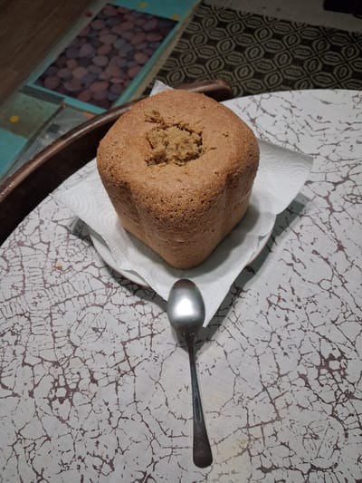 An overhead, eye-level, medium shot shows a freshly baked loaf of bread centered on a white paper napkin, which is on a white plate with a faint red rim. The bread is square-shaped with a golden-brown crust and a slightly sunken, rough top where it appears to have been disturbed or broken open. A silver spoon rests diagonally in front of the bread, with its bowl facing the loaf. The bread and spoon are positioned on a white tablecloth with a dark brown, crackle-like pattern. In the background, out of focus, is a dark brown wooden tray with a textured blue surface containing a mosaic of smooth, multi-colored stones. To the right of the tray, a dark fabric with a geometric pattern is visible. The overall scene suggests a domestic setting, possibly a kitchen or dining area, in Goran, Bulgaria.