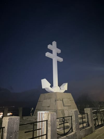 The image shows the Free French Memorial in Gourock, United Kingdom. A white Cross of Lorraine, the symbol of the Free French Forces during World War II, is mounted on a stone plinth. The sky is a deep blue, suggestive of nighttime. To the right of the memorial are concrete structures with black metal railings.