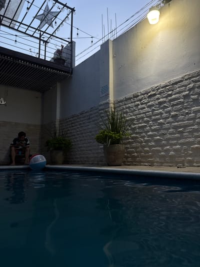The scene is outdoors, alongside a swimming pool filled with dark-colored water. A person is sitting at the edge of the pool. Near the person is a red, white, and blue beach ball. Adjacent to the pool is a textured, light-colored brick wall with two potted plants affixed to it. Above the wall is a portion of a building with a light fixture and what appears to be a balcony with decorative lights and a star. The image is taken from an unusual angle, giving a tilted perspective. The location is Chiapa de Corzo, Mexico.
