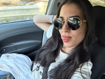 A woman with long, dark hair is reclining in what appears to be the back seat of a car. She is wearing round sunglasses with gold frames and is sticking her tongue out slightly. She is wearing a light-colored t-shirt and has a smartwatch or fitness tracker on her wrist. The car seems to be moving, as indicated by the blurred scenery visible through the window. There is a train track seen through the window with the background being of vegetation and other tracks. The city of Komarevo, Bulgaria cannot be confirmed from this image.