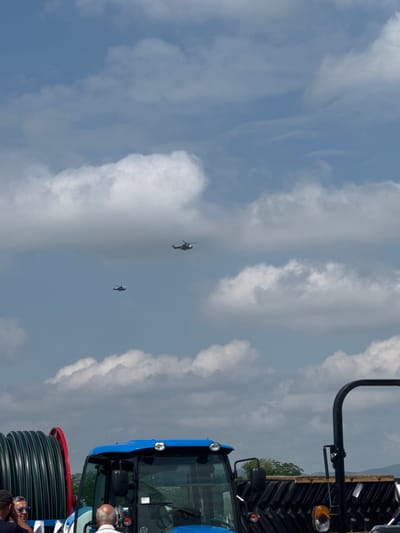 The image, oriented correctly, captures an outdoor scene under a bright, partly cloudy sky, likely during midday or early afternoon. The sky, a mix of blue and scattered white clouds, occupies most of the frame.
Two individuals are visible flying personal aerial devices in the mid-ground. These devices resemble small, open-cockpit helicopters or advanced jetpacks, each with a person piloting it. One flyer is positioned higher and further away, while the other is lower and closer to the viewer, both silhouetted against the sky.
In the bottom right foreground, the front section of a blue tractor, likely a New Holland model, is prominent. Features like its cabin, a headlight, and a large black roll bar extending upwards are visible. Adjacent to the tractor, a large, black industrial spool of coiled tubing or hose is also present.
Several people are partially visible in the bottom right, near the tractor and the spooled tubing. One man with a bald head and a white t-shirt is clearly looking upwards, seemingly observing the flying devices. Another person wearing sunglasses is also present.
The scene appears to be an outdoor event or demonstration, possibly combining an air display with an agricultural or machinery exhibit, set against a backdrop of distant hills and scattered trees. The location is Madzherito, Bulgaria. No discernible text is visible on any of the subjects.