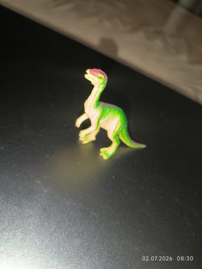 A miniature, brightly colored toy dinosaur, predominantly green with pink accents on its head and tail, is positioned on a dark, reflective surface. The toy is depicted from a side profile, with its head tilted upwards. The background shows a blurred diagonal line, suggesting a surface change, with a lighter, textured area above it. A timestamp in the bottom right corner reads "02.07.2026 08:30". There is no discernible location in Kattasu, Uzbekistan, or any human presence depicted in the image.