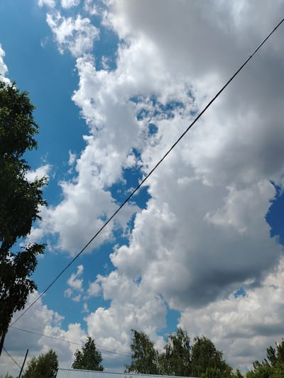 The image captures a view of the sky and treetops in Жернов ауылдық округі, Kazakhstan. A thick, dark power line stretches diagonally across the frame from the upper left to the lower right. The sky is a mix of bright blue patches and large, fluffy white and grey clouds, suggesting a partly cloudy day. On the left side of the image, the dark green foliage of a tree is visible, extending into the frame from the top. Along the bottom, the tops of several deciduous trees with green leaves are seen, and a silver corrugated metal roof is partially visible below them. The scene appears to be outdoors, possibly in a rural or suburban area, given the presence of trees and overhead wires.