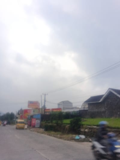 The image depicts a blurred street scene in what is identified as the city of Morrumbala, Mozambique, under an overcast sky. A paved road occupies the lower portion of the frame. In the immediate foreground on the right, a person wearing a blue helmet is operating a light-colored motorcycle, exhibiting motion blur. Further up the road on the left, a yellow vehicle is present, either stationary or moving slowly. Behind the yellow vehicle, additional indistinct vehicles and figures are discernible.

On the left side of the road, a large billboard with red and white signage is visible above smaller, multi-colored structures, possibly commercial establishments. These structures are partially obscured by vegetation. On the right side of the road, a strip of green terrain separates the roadway from a building featuring a dark, pitched roof and dark-toned walls. Behind this structure, another lighter-colored, multi-story building is visible. Multiple utility poles and overhead power lines traverse the mid-ground. The sky is uniformly light grey and overcast, dominating the upper half of the image.