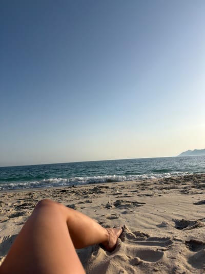 The image depicts a serene beach scene in Grândola, Portugal, bathed in the soft light of what appears to be late afternoon. The perspective is from the point of view of someone relaxing on the sand, with their legs and feet visible in the foreground. The gentle waves of the Atlantic Ocean lap at the sandy shore, leaving a foamy white line where the water meets the land. The sand is imprinted with numerous footprints, suggesting a day of activity.
In the distance, the hazy outline of a coastline or distant hills is visible against a pale, clear sky that transitions to a brighter hue near the horizon. The overall atmosphere is one of tranquility and leisure, evoking a sense of peace and relaxation. There are no visible signs or text in the image. The weather seems to be pleasant, with clear skies and calm seas.