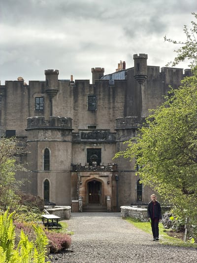 The image captures a gray stone castle, likely Dunvegan Castle in Dunvegan, United Kingdom, under a cloudy sky. The castle's architecture features multiple levels, windows, and turret-like structures. A gravel path leads towards the castle's entrance. A person wearing dark clothing stands on the left side of the path, near some green foliage. Vegetation, including ferns and trees with budding leaves, flanks the path.